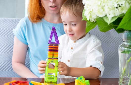 Caucasian boy is sitting with his mother having fun playing with magnetic constructor. Selective focus. Early development in kindergarten and at home.